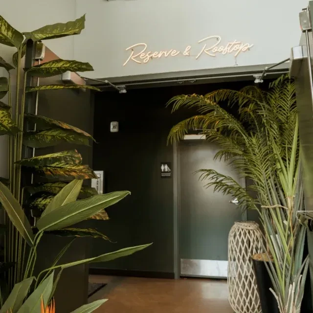 Interior hallway with green plants, a dark door, and a 'Reserve & Rooftop' neon sign.