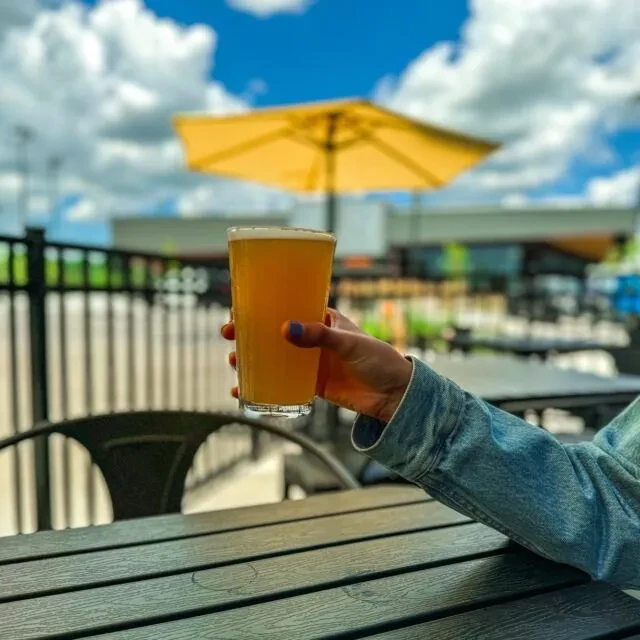 Hand with purple nails holds foamy beer on a sunny outdoor patio.
