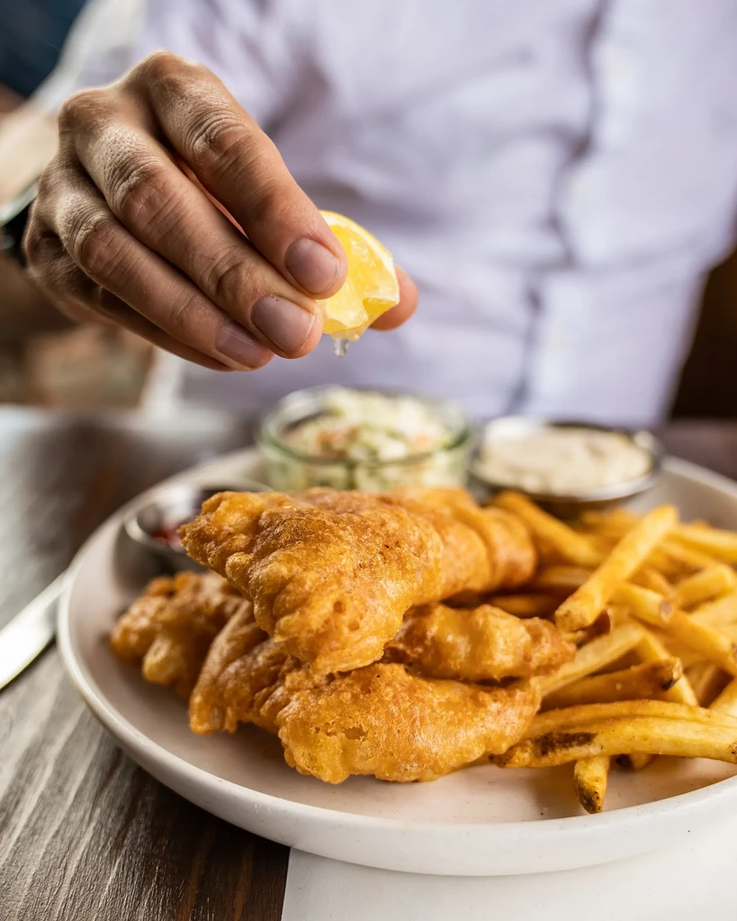 Close-up: Hand squeezing lemon over golden fish and chips.