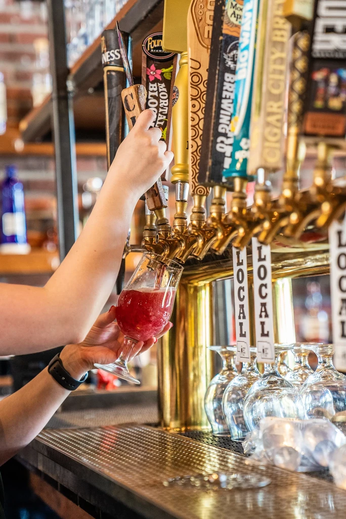 Hands pour frothy red drink from a Mortal Blossom tap into a glass.