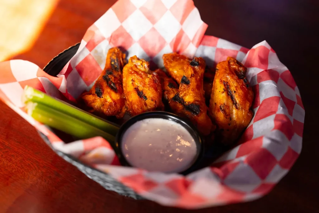 Charred buffalo wings in a basket with celery and dipping sauce.