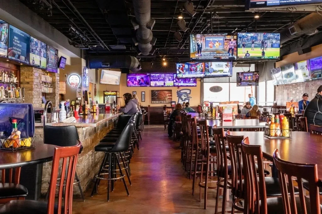 Sports bar interior with long bar, tables, and numerous TVs.