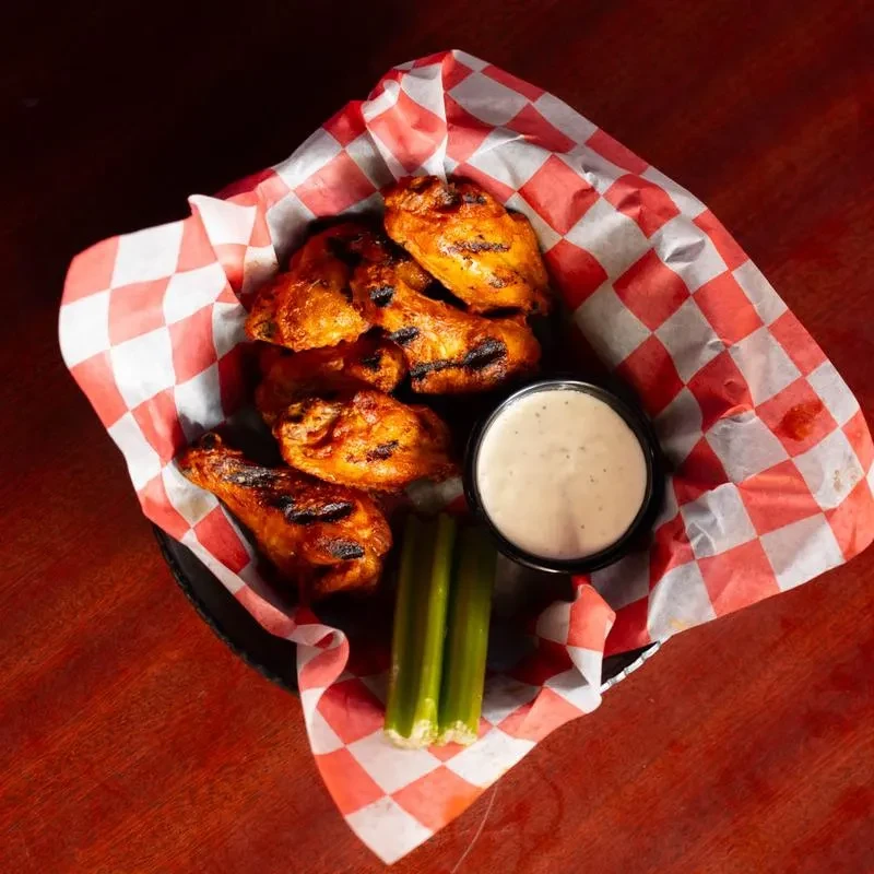 Grilled chicken wings, dipping sauce, and celery in a checkered basket.