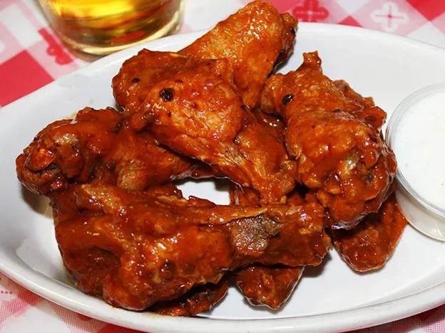 Piled sauced chicken wings, dipping sauce, and beer on red checkered tablecloth.