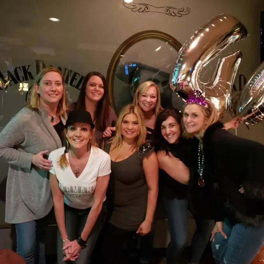 Seven women celebrate a birthday in a dim bar with balloons and crown.