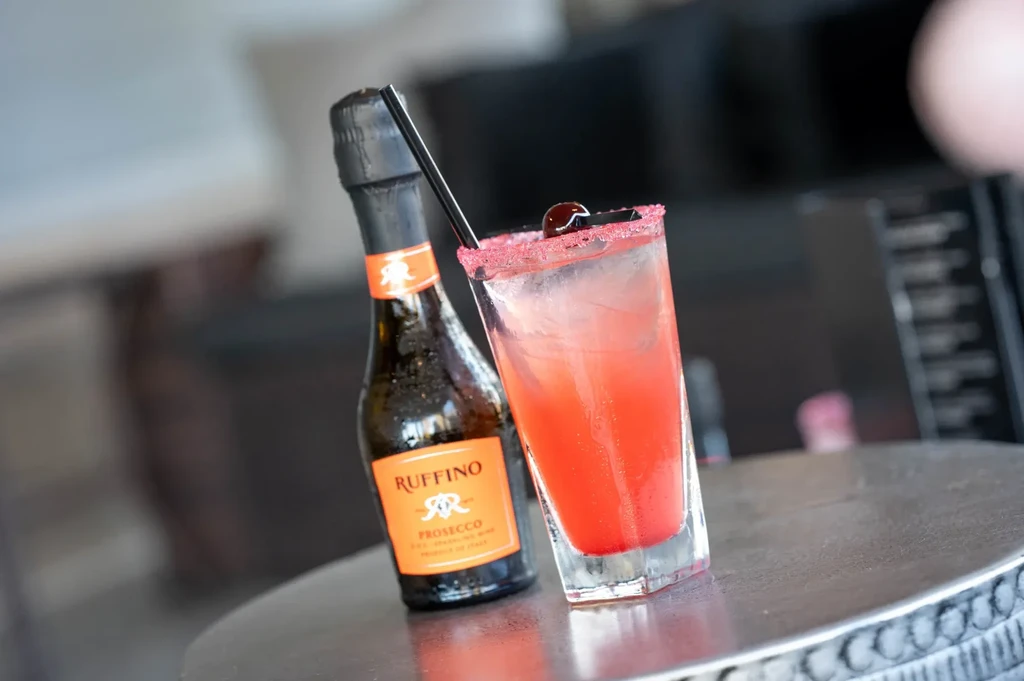 Prosecco bottle and pink cocktail with cherry on a metal table.