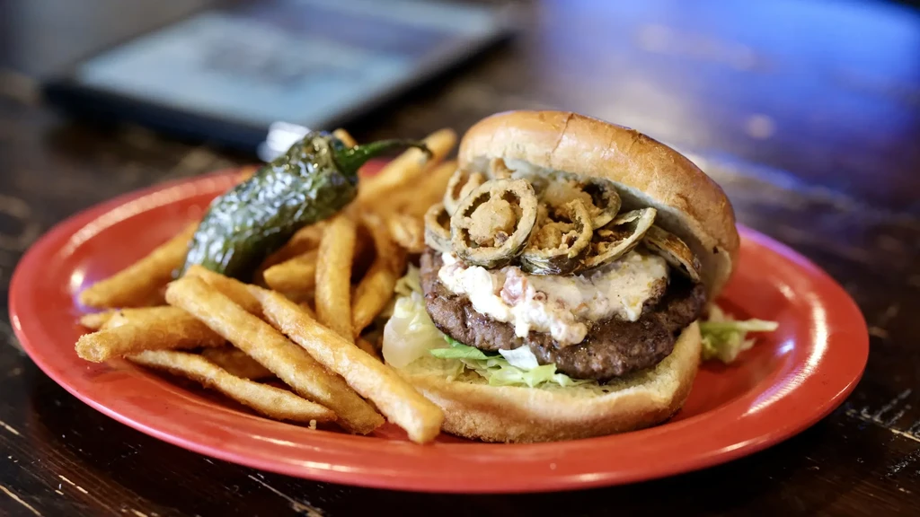Gourmet hamburger with jalapeños, fries, and a pepper on a red plate.