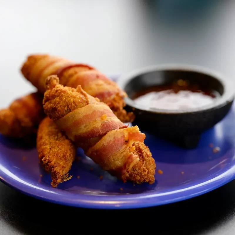 Three crispy bacon-wrapped breaded tenders with dipping sauce on blue plate.