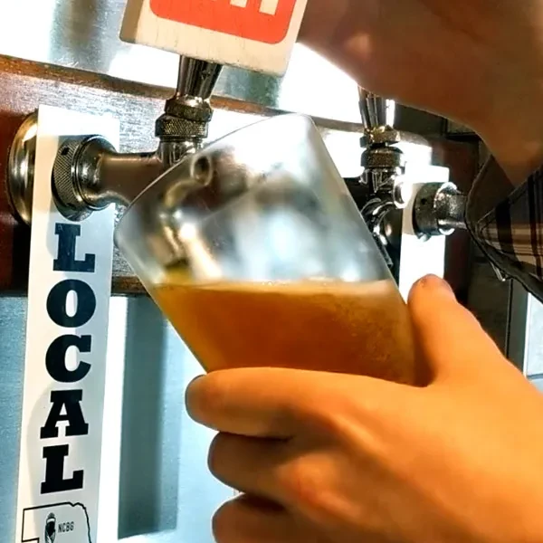 Hands pour frothy amber beer from a tap with "LOCAL" handle.