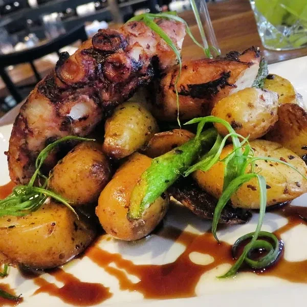 Plated grilled octopus, roasted potatoes, green herbs, and dark sauce.
