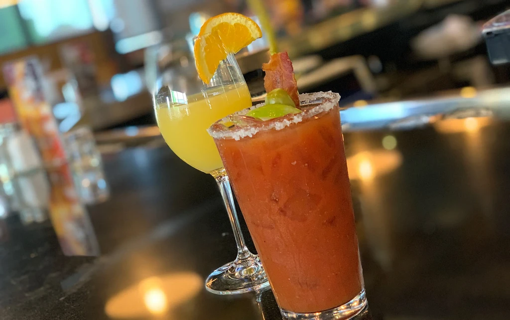 Mimosa with orange and Bloody Mary with bacon on dark surface.