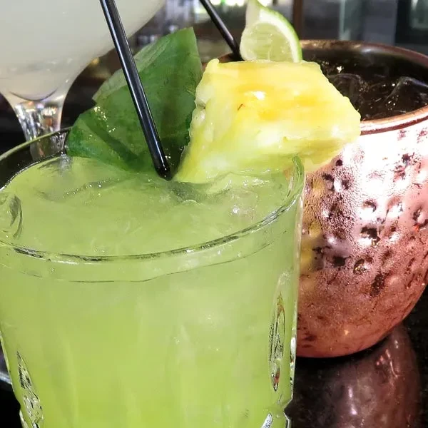 Green cocktail with pineapple, Moscow Mule, and martini on dark surface.