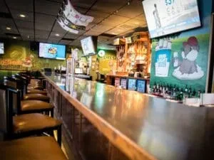 Dim bar interior with reflective counter, empty stools, bottles, and TVs.