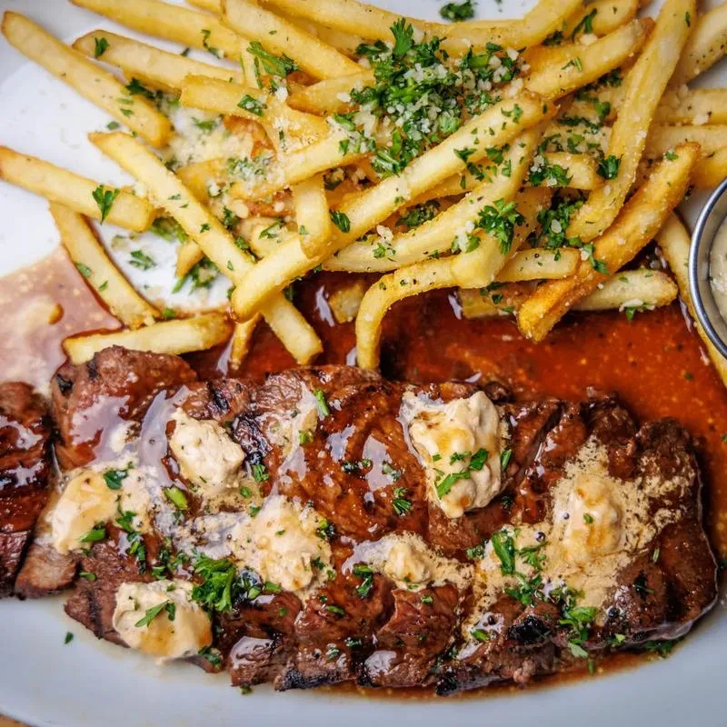 Sliced steak with sauce, fries with cheese, parsley, and dipping sauce.