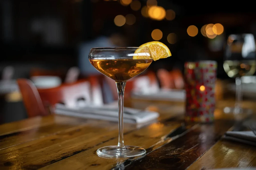 Amber cocktail in coupe glass, orange slice, rustic table, blurred background.
