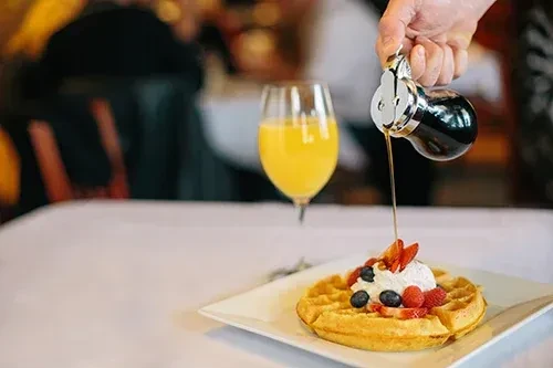 Close-up of golden waffle with berries, whipped cream, and syrup being poured.