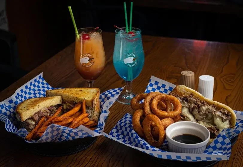 Two sandwiches with fries, onion rings, and colorful Goose Island cocktails.