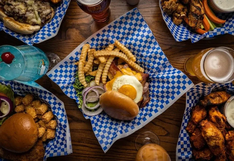 Overhead shot: burgers, cheesesteak, wings, fries, tots, beers, blue cocktail.