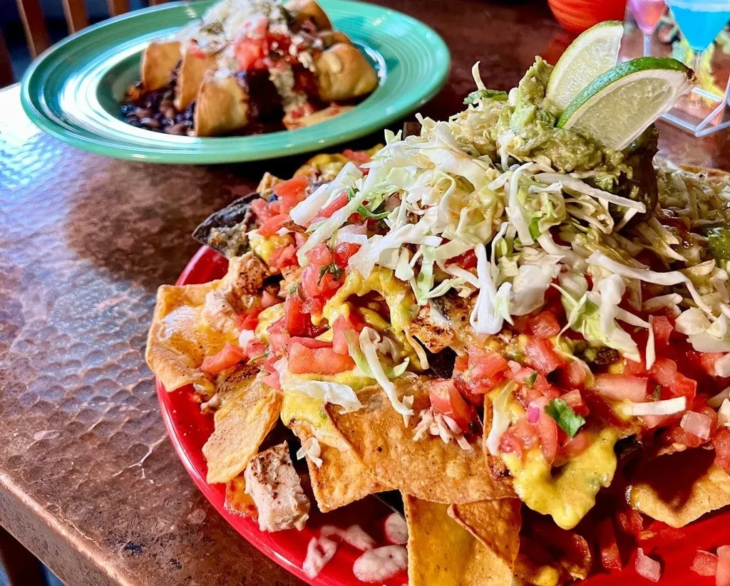Large nachos with chicken, guacamole, and a blurred meal and drinks.