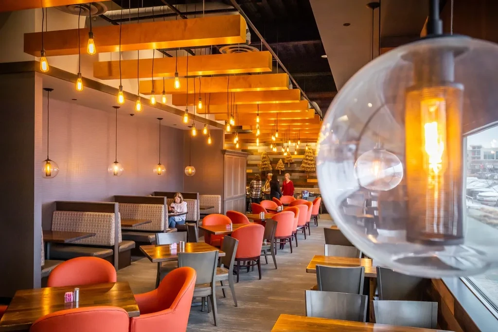 Restaurant interior featuring booth seating, orange chairs, and hanging Edison bulbs.