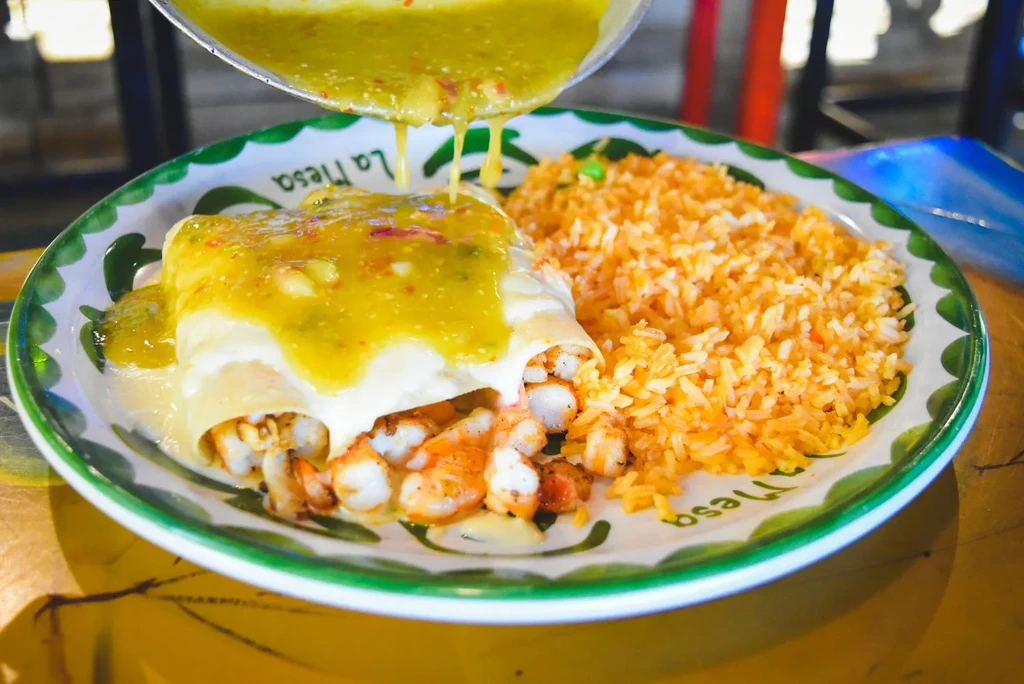 Shrimp enchiladas with green sauce being poured, served with rice.
