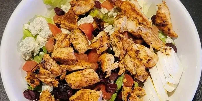 Chicken salad in white bowl with lettuce, tomatoes, cheese, dried fruit.
