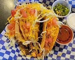 Hard-shell tacos with meat, cheese, and three condiments on checkered paper.