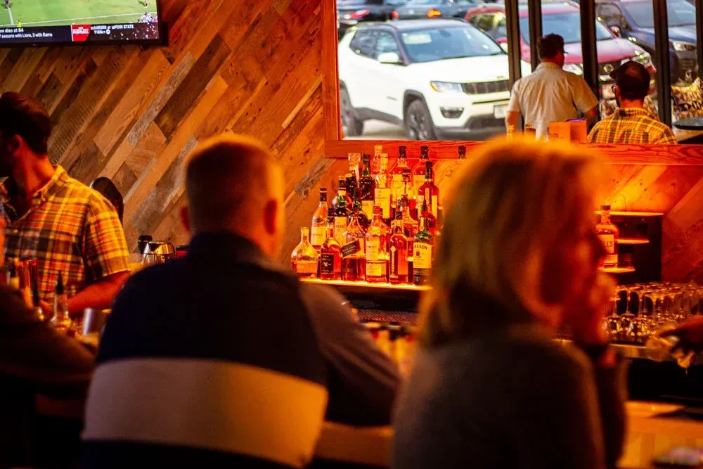 Warmly lit bar with illuminated liquor bottles, patrons, and a TV.