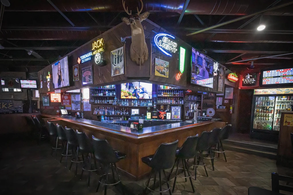 Dimly lit bar with liquor, neon signs, multiple TVs, and deer head.