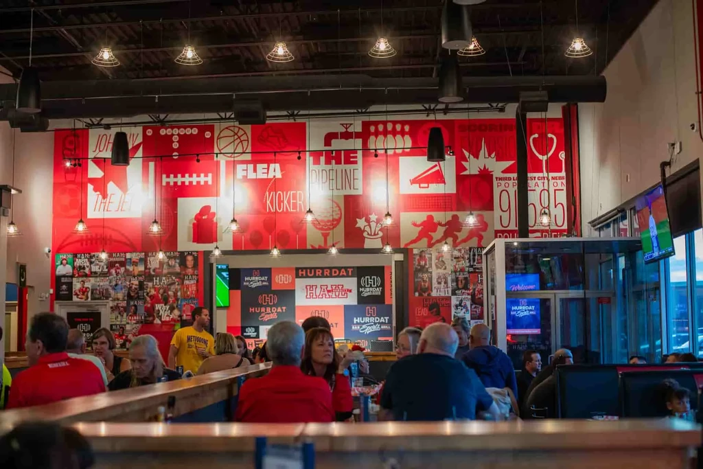 Bustling industrial-style sports bar with sports murals and many patrons.