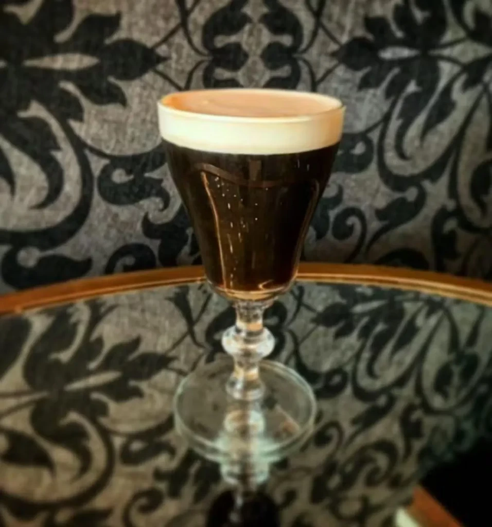 Stemmed glass with dark foamy drink on reflective surface, ornate background.