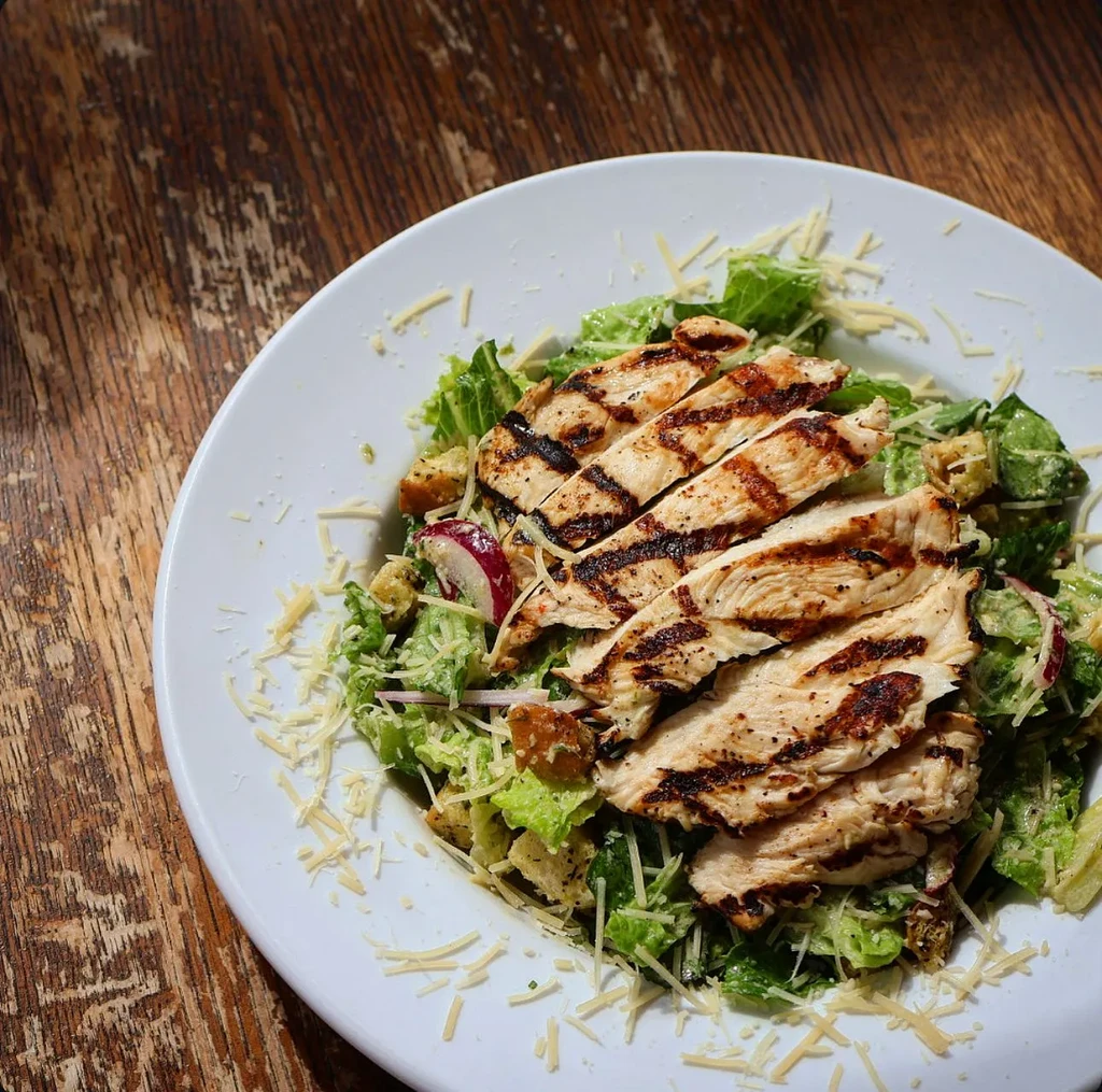 Overhead grilled chicken Caesar salad on a rustic wooden table.