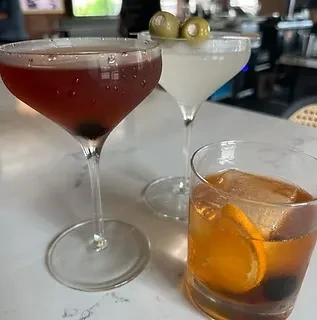 Three cocktails: purple coupe, clear martini, amber rocks glass on bar.