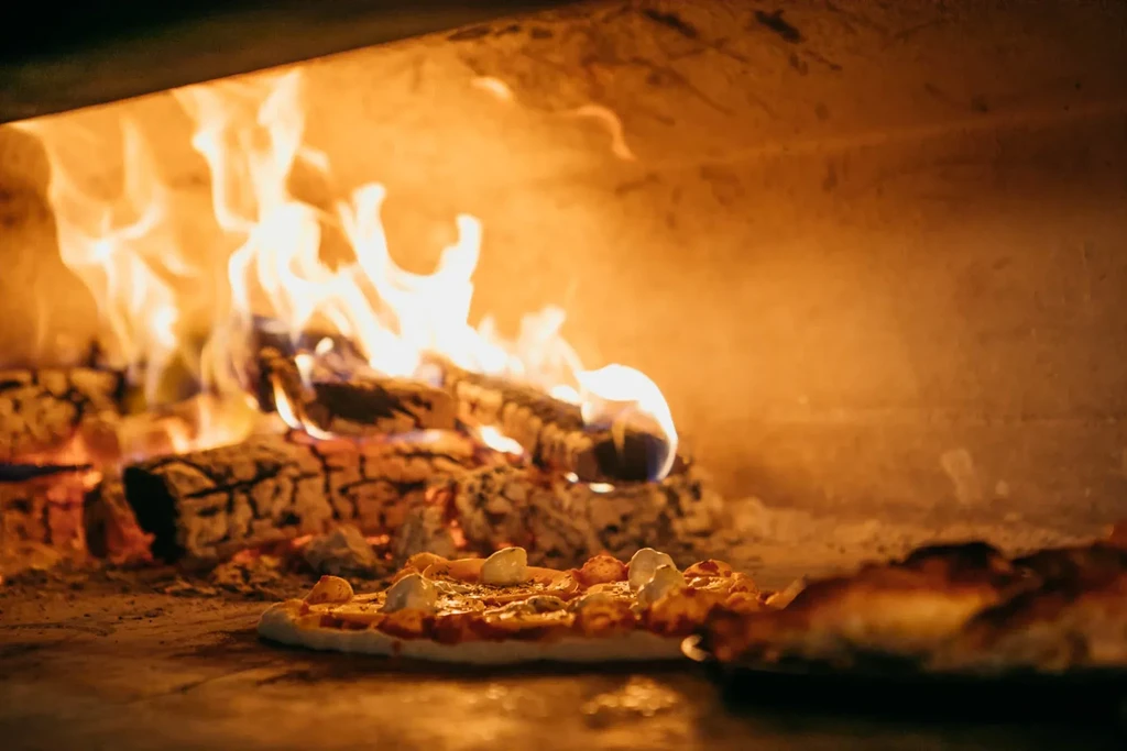 Wood-fired oven interior with a pizza baking next to glowing flames.