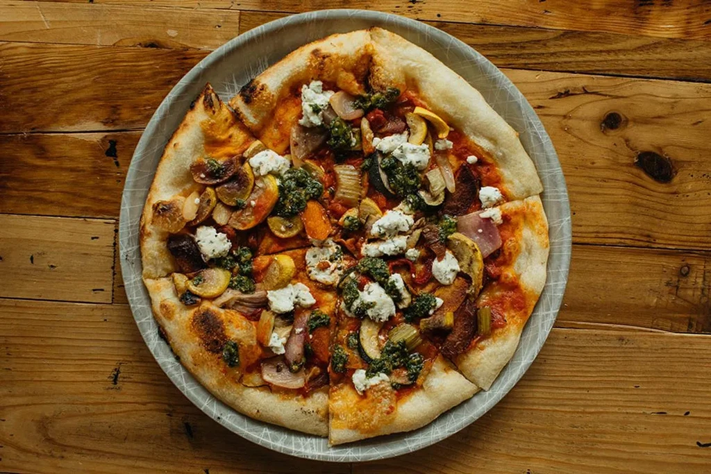 Wood-fired pizza with vegetables, cheeses, and pesto on a rustic table.