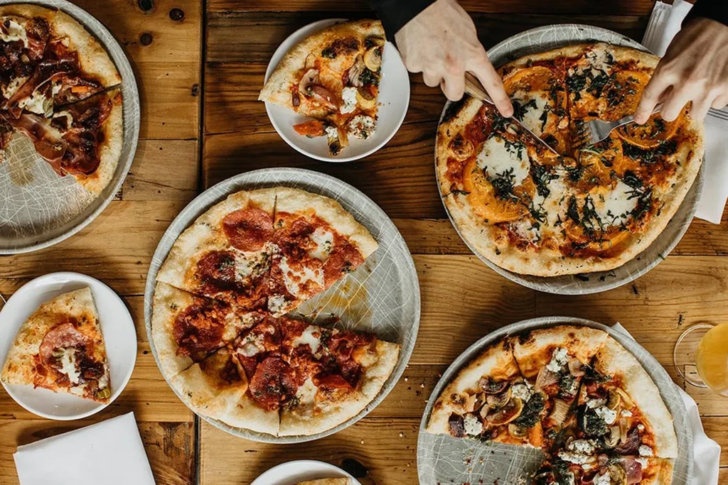 Four varied pizzas on a wooden table, hands cutting a slice.