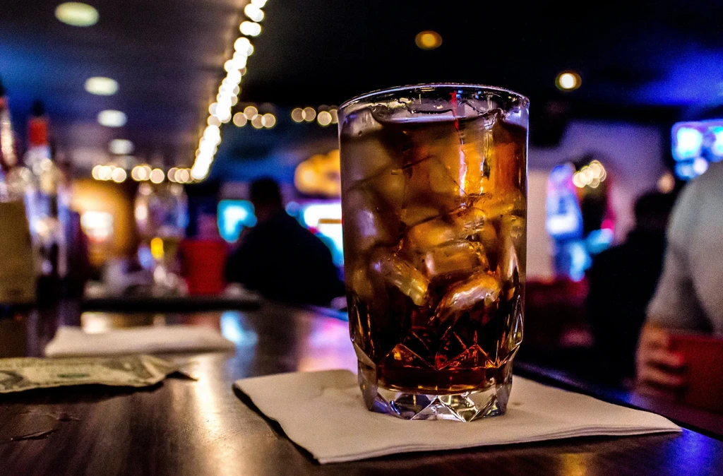 Amber drink with ice on bar, dollar bill, blurred lively background.