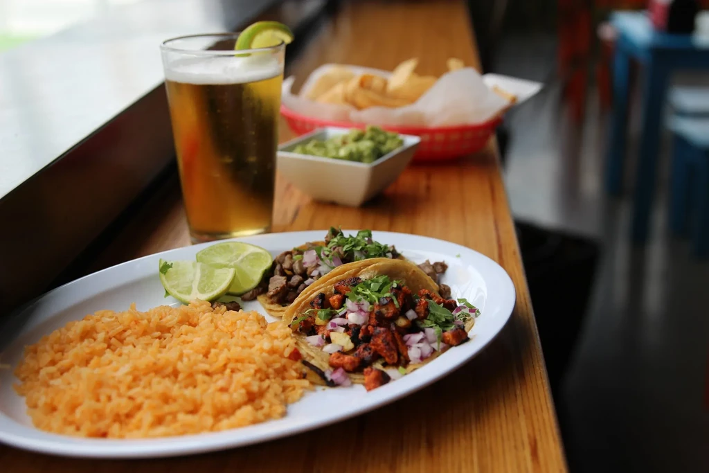 Plate of tacos with rice, beer, guacamole, and chips on wood.