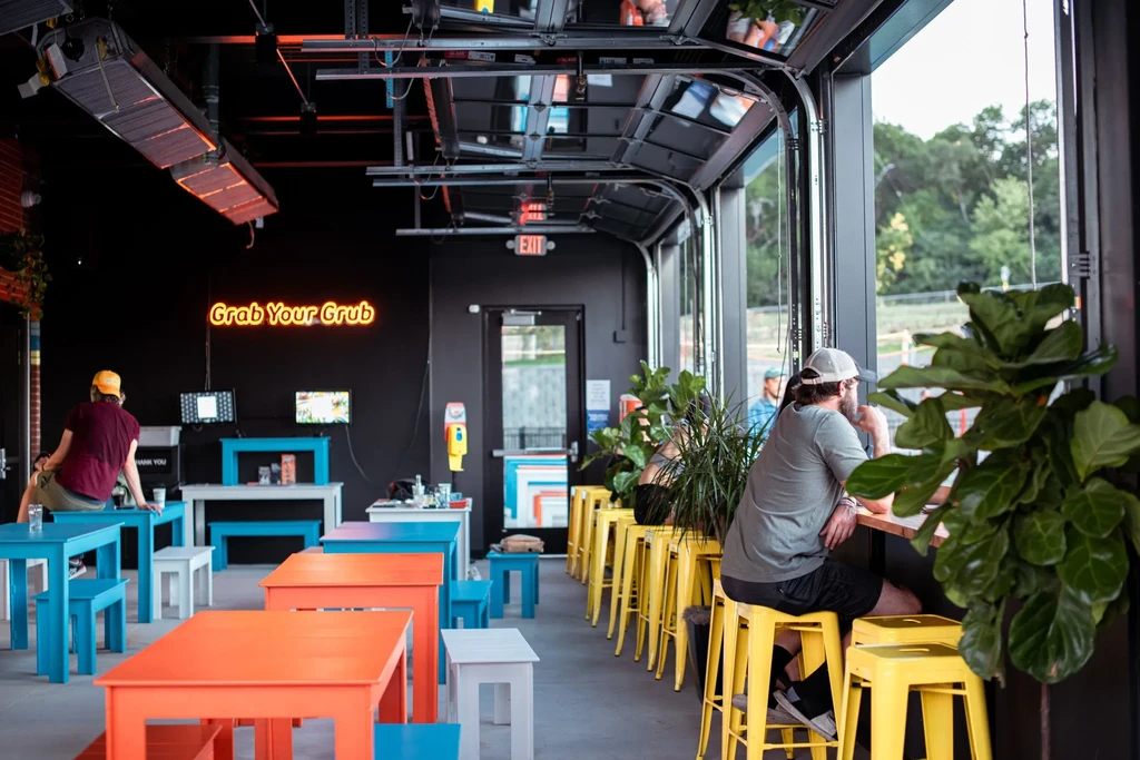 Modern industrial-chic restaurant interior with colorful seating and neon sign.