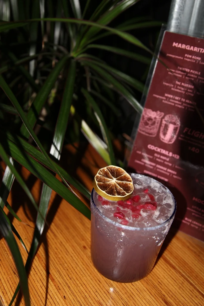 Colorful purple cocktail with salted rim, lime, and pomegranate on wood.
