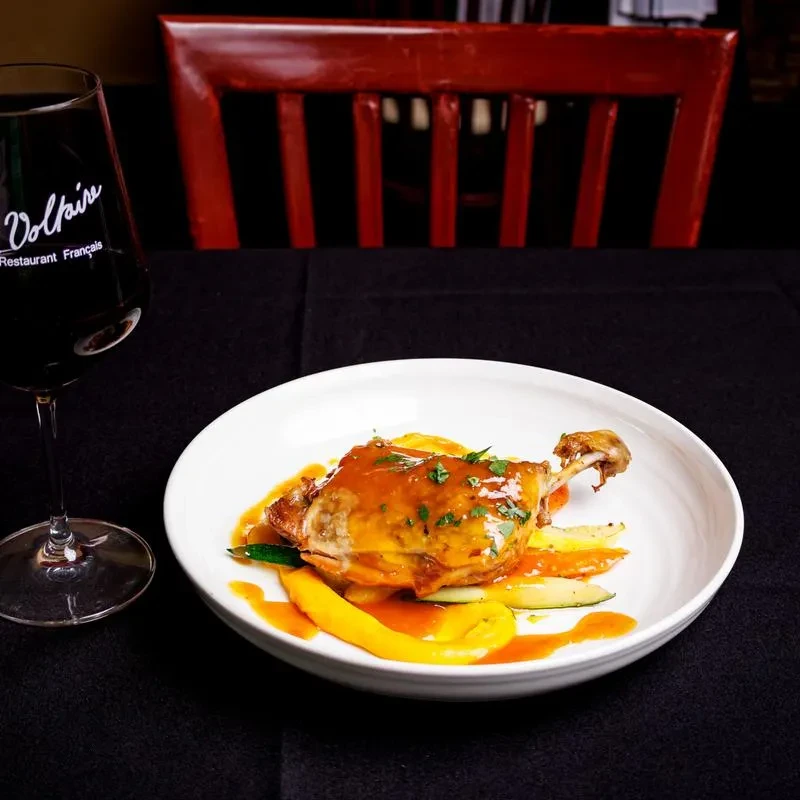 Plated poultry dish with sauce, vegetables, and red wine at Voltaire Restaurant.
