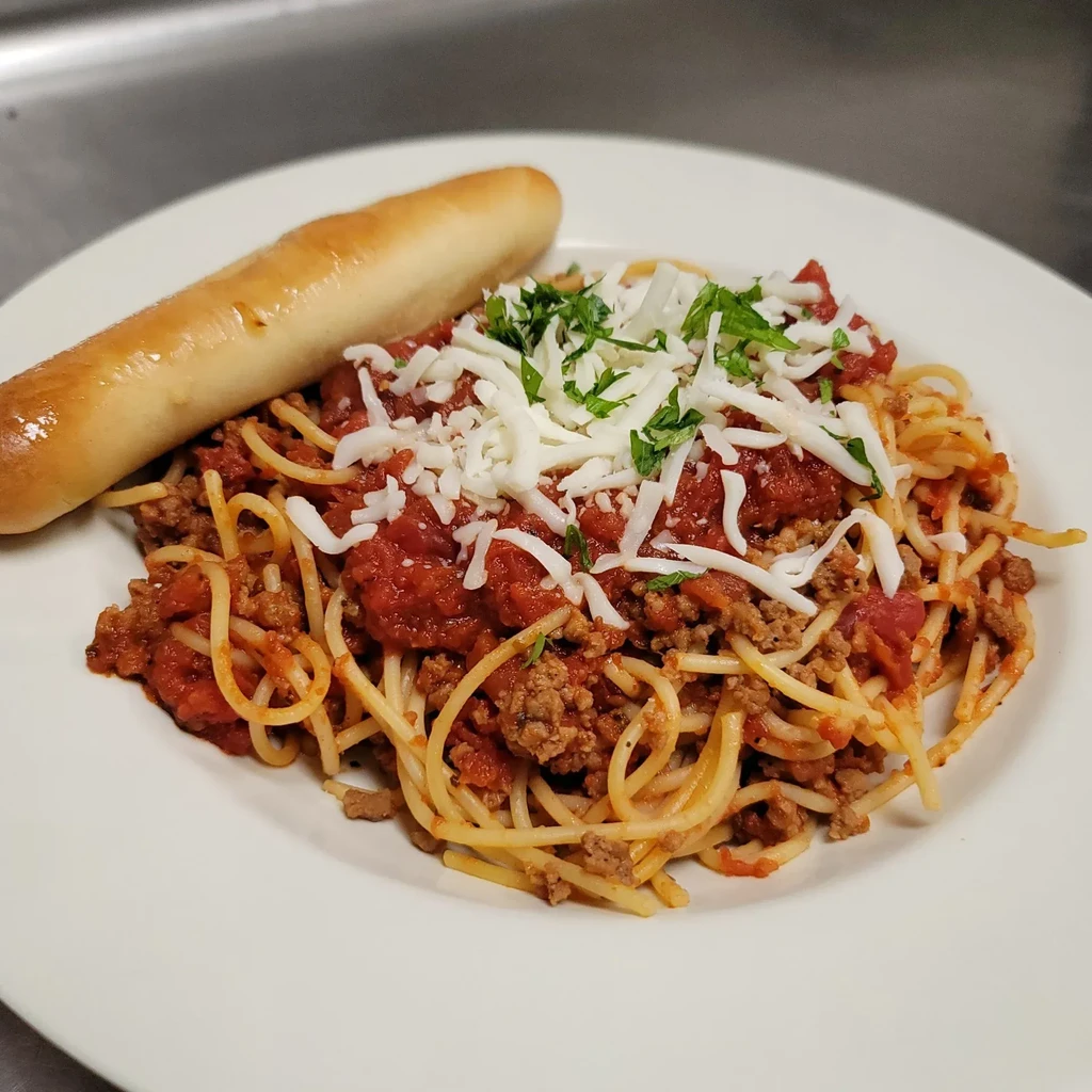 White plate of spaghetti with meat sauce, shredded cheese, green herbs, breadstick.