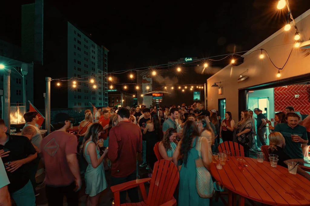 Nighttime outdoor bar scene with people, string lights, and city skyline.