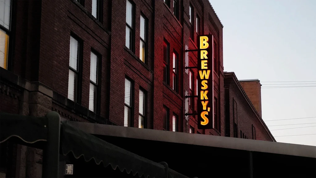 Vertical glowing BREWSKY'S neon sign on a red brick building at dusk.
