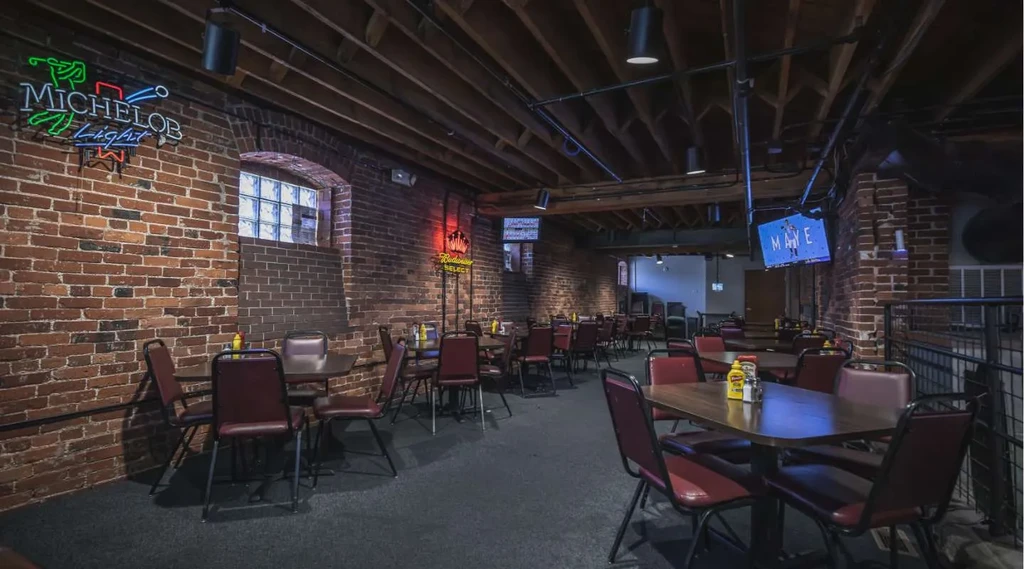 Dimly lit rustic bar interior with exposed brick, tables, and neon signs.