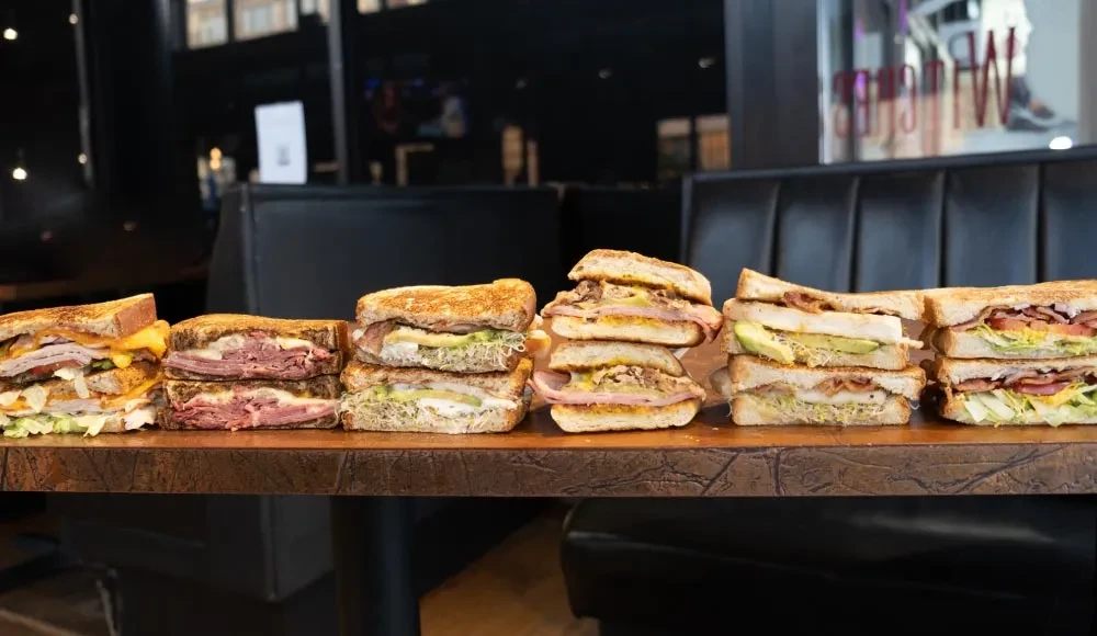 Six distinct large, stacked sandwiches on a rustic wooden surface.