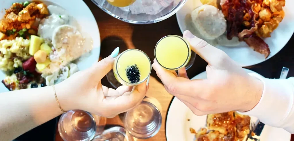 Hands toast yellow drinks, one with dark pearls, above food.
