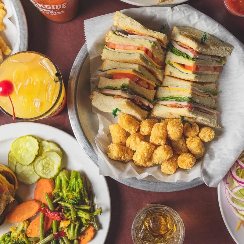Club sandwiches, tater tots, pickles, vegetables, and drinks on a table.