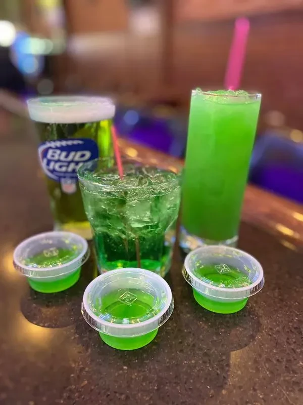 Green drinks, Bud Light cup, and shots on a dark bar.