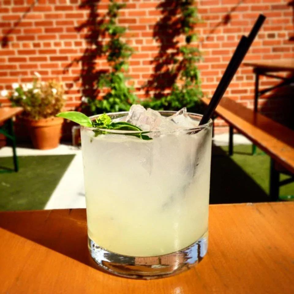 Yellow-green cocktail with ice and green sprig on a sunny wooden table.